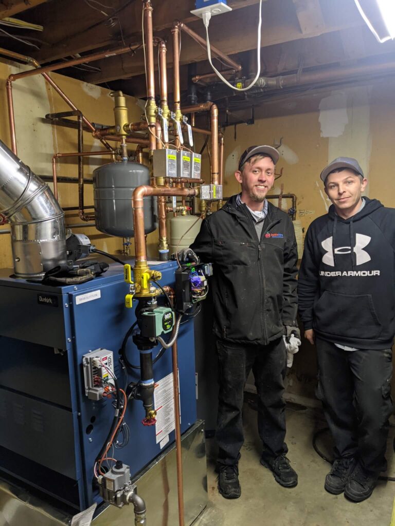 two apple aire technicians standing in front of a newly replaced boiler. In the basement with piping and blue boiler